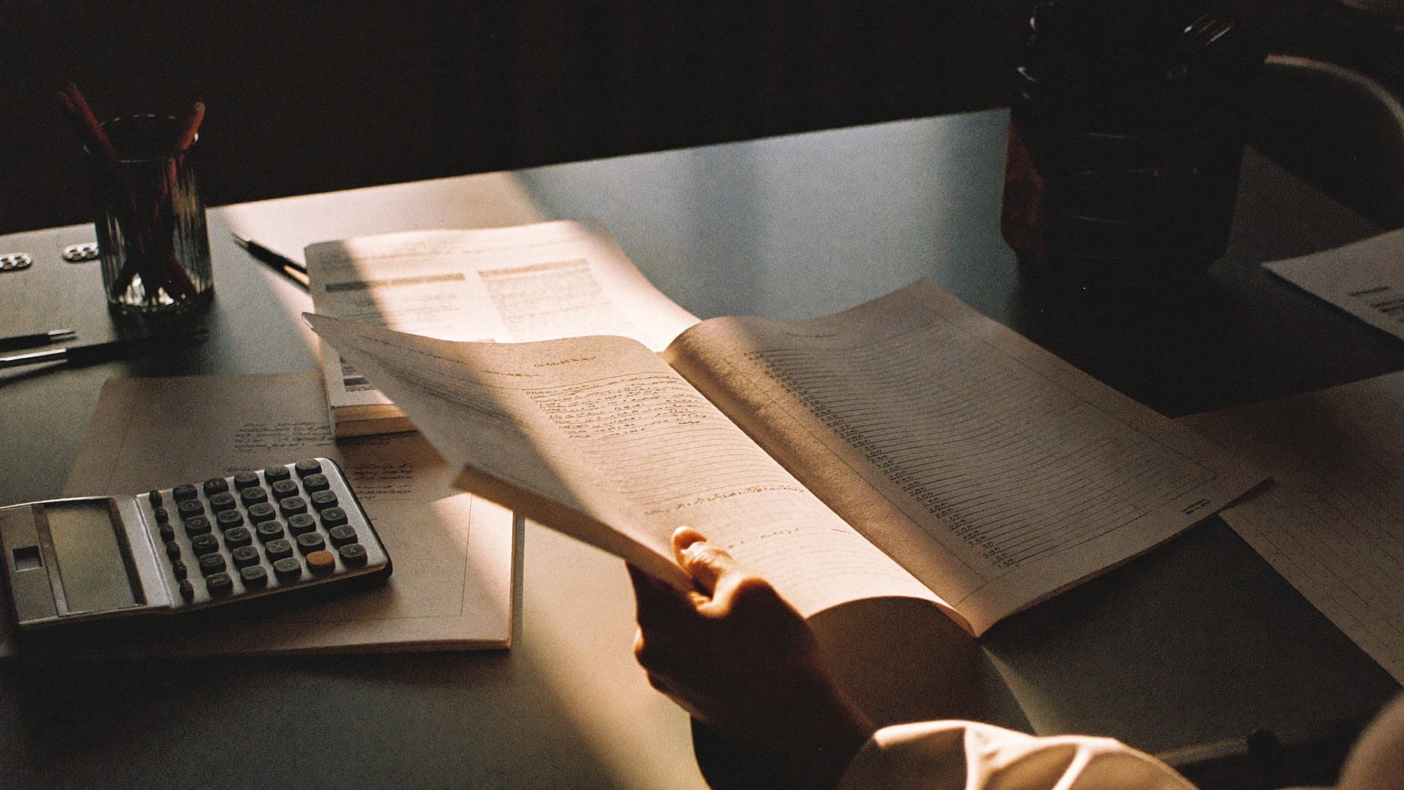 Mesa cheia de planilhas impressas e calculadora sob luz fria, uma mão folheando páginas